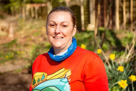 Smiling person wearing a blue scarf and a red tshirt with daffodils and trees in the background