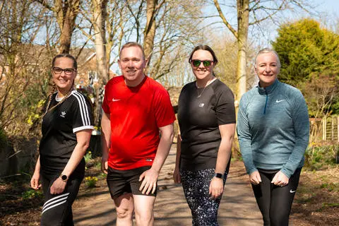 Four smiling people dressed in sportswear stood on a countryside lane surrounded by trees