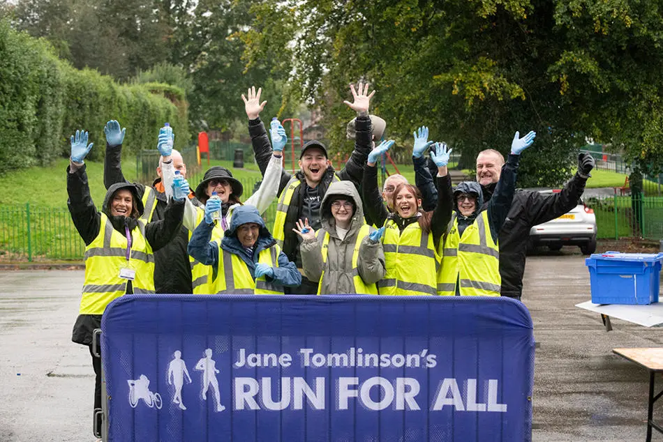 A group of smiling people wearing hi-viz vests standing in front of a banner for Jane Tomlinson's Run for All