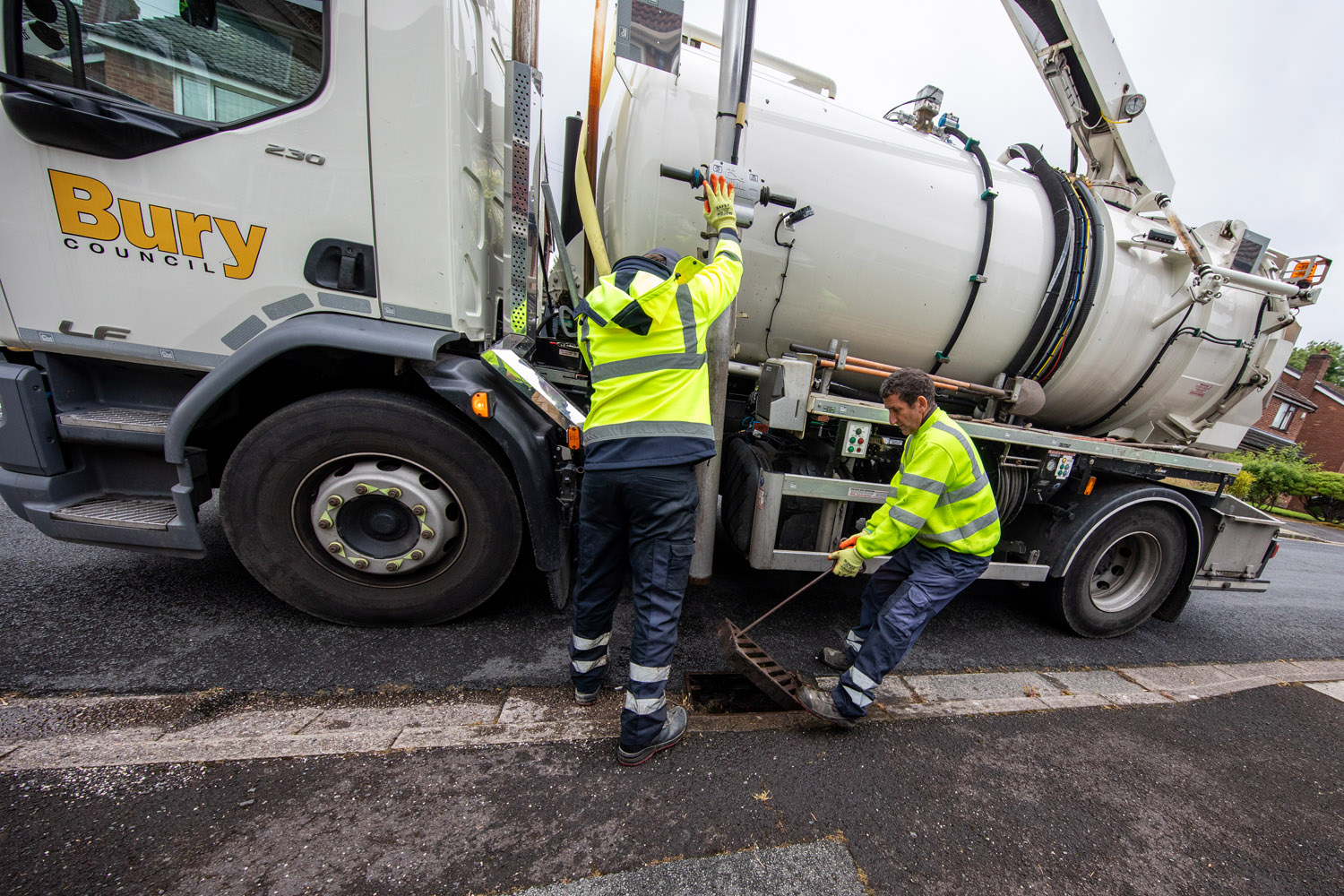 Two people stood in front of a large vehicle, one person is pressing a button on the vehicle and one person is lifting a drain cover