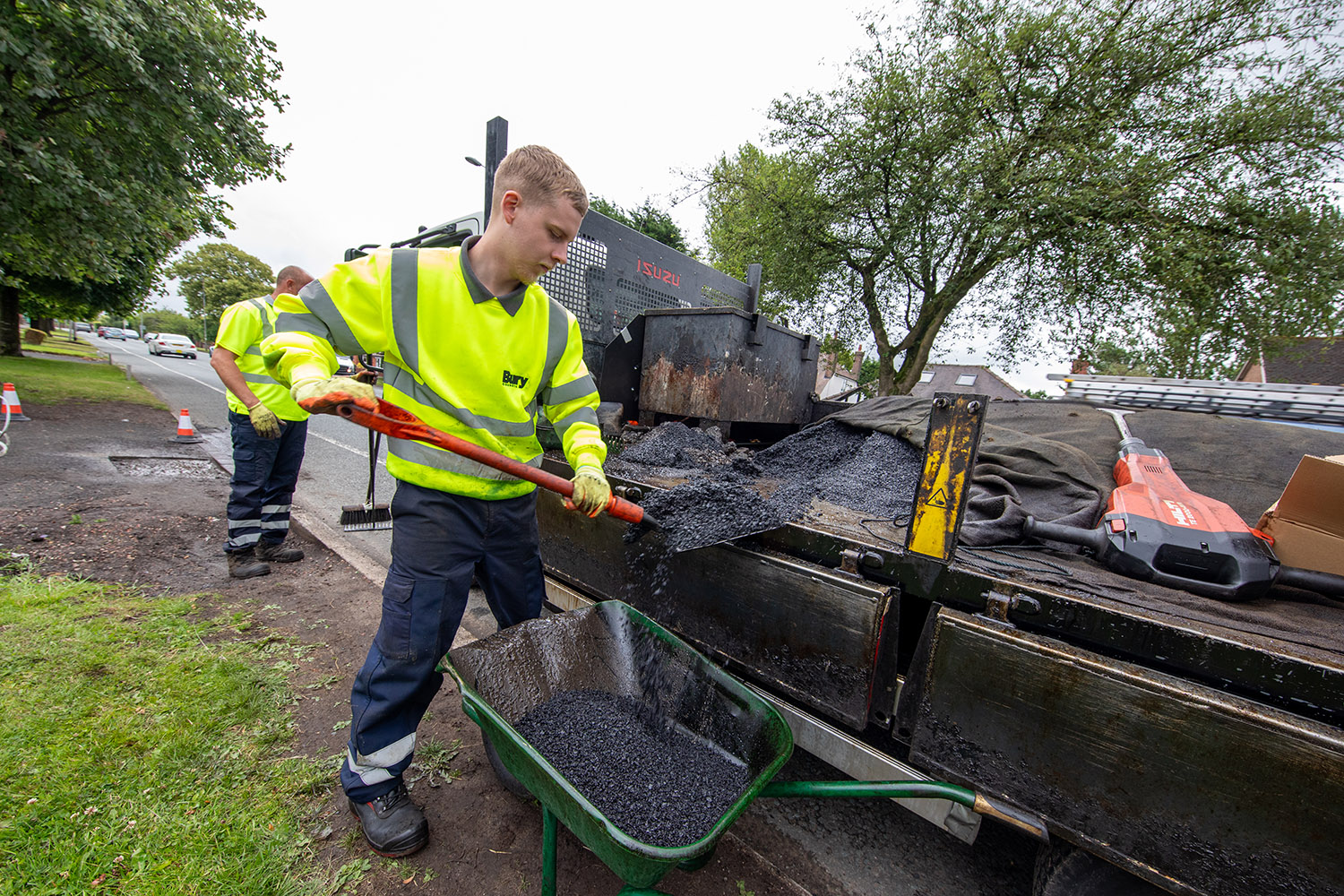 A person in high visibility clothing using a spade to collect tarmac from the back of a large truck