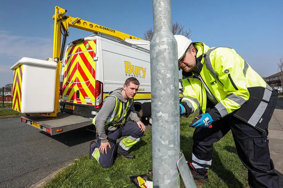 A person in high-visibility clothing working on a streetlight column which has its cover removed