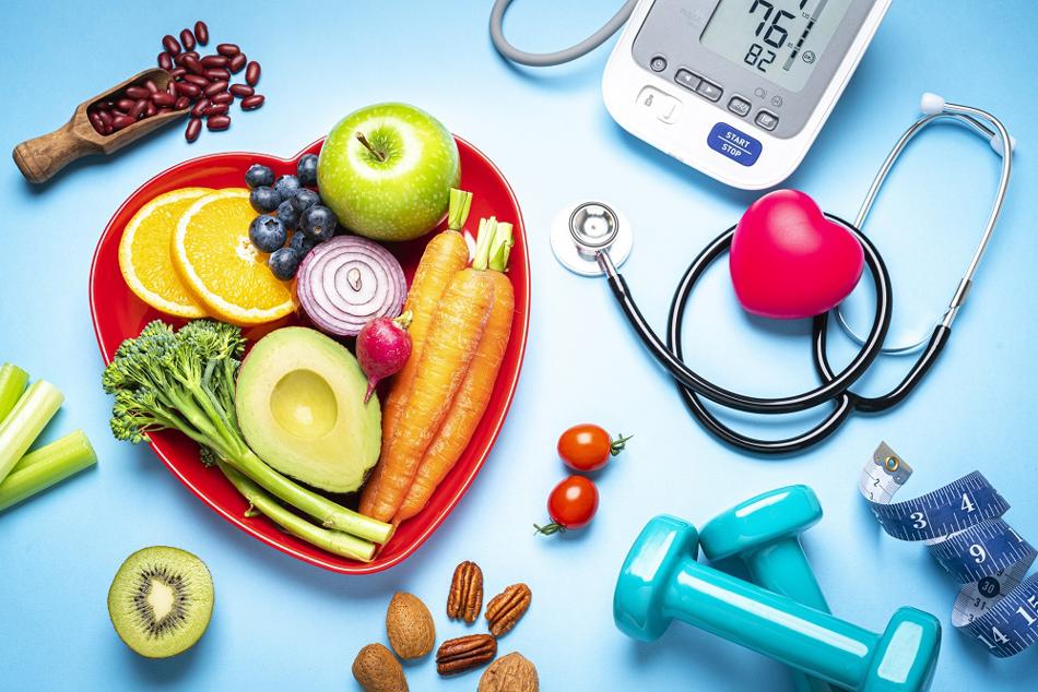 Heart shaped bowl of fruit and vegetables next to some weights and a stethoscope.