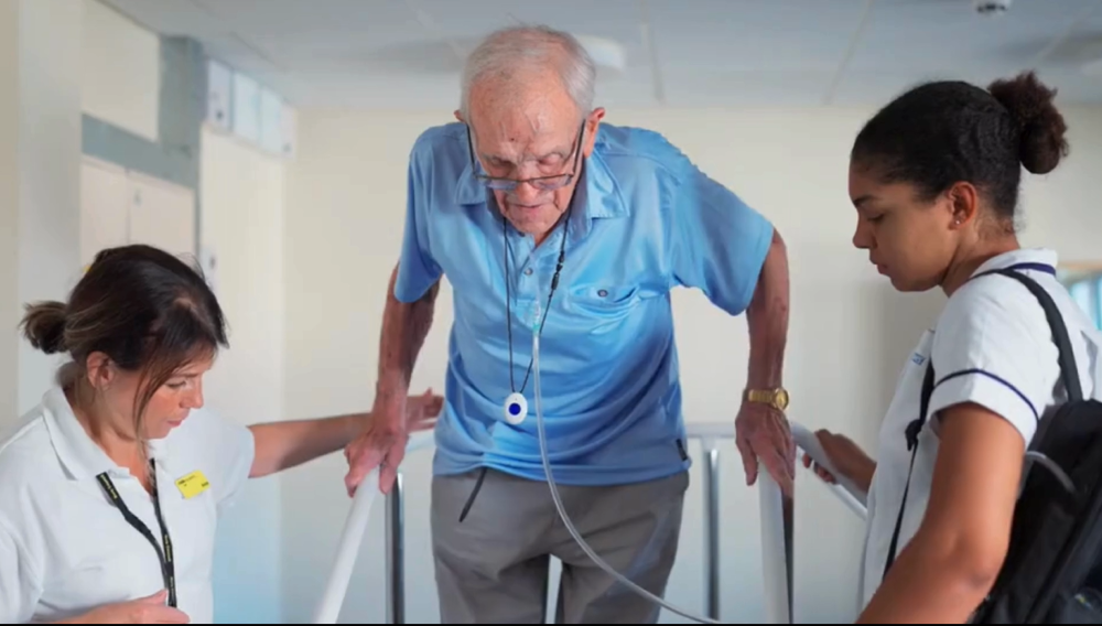 Two nurses helping an older person on a walking frame.