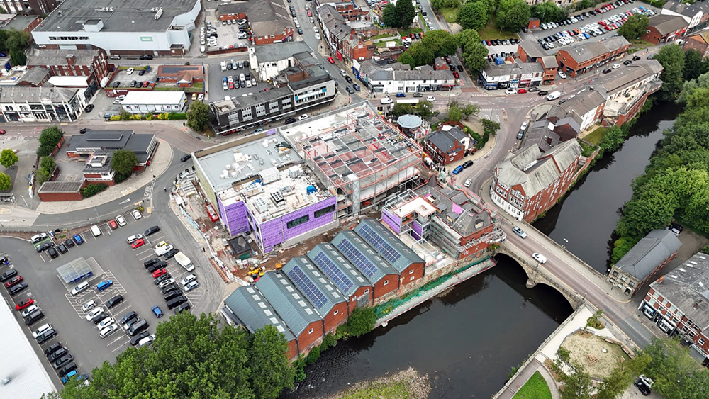 A large building on a construction site alongside a river and surrounded by a road and buildings