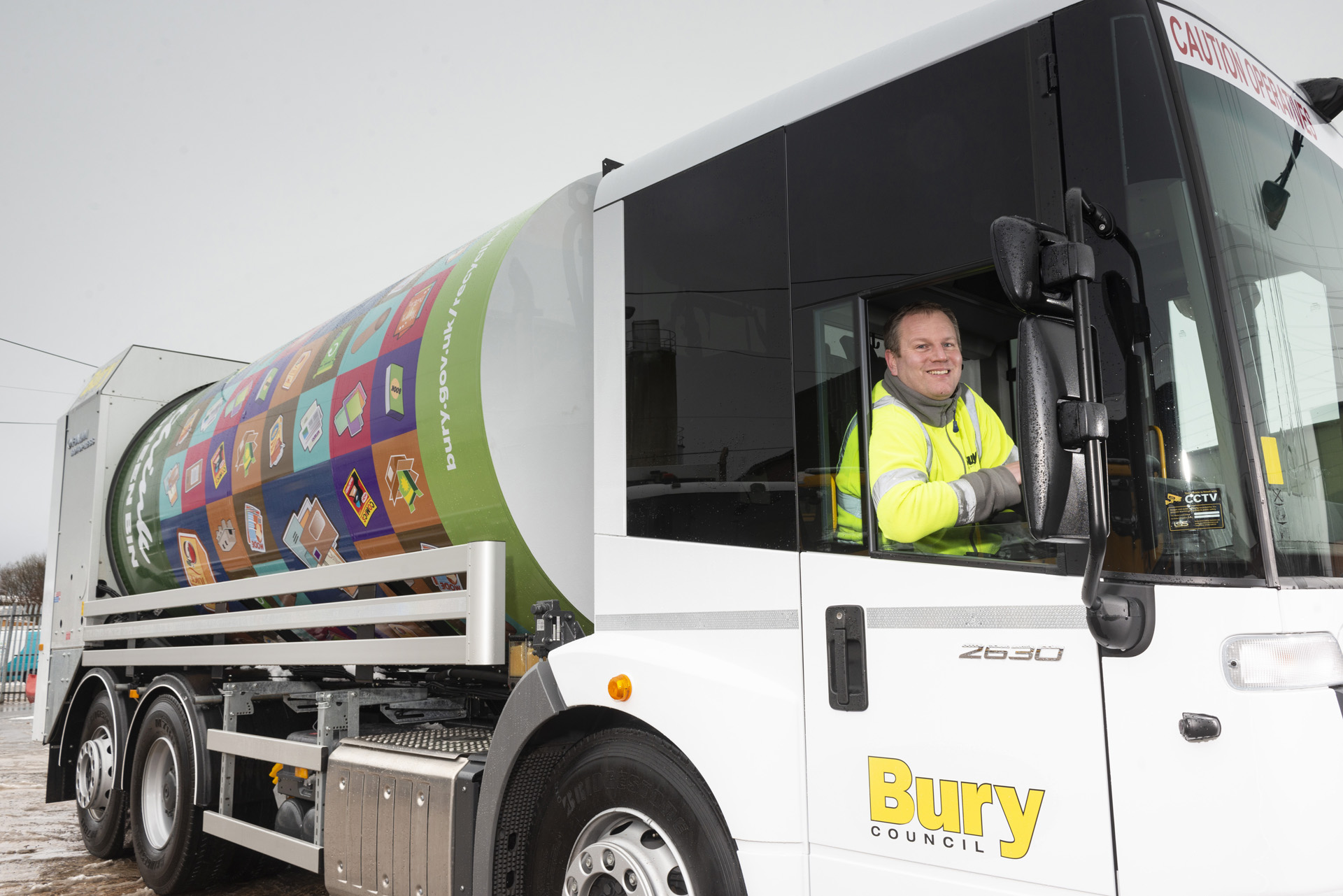A side on view of a bin collection truck with a driver in the cab
