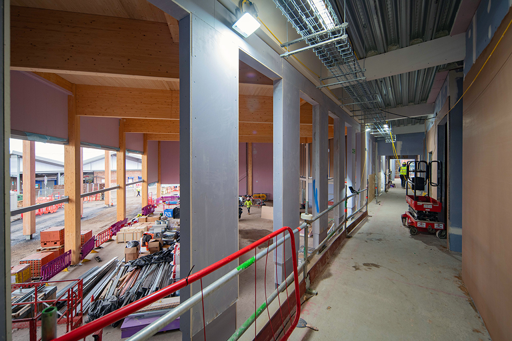 The inside of a large building during the construction phase. The building has a wooden ceiling, large windows along one side and an open gallery on the first floor.
