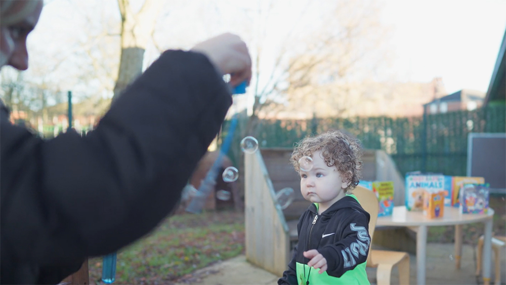 Young child stood in a playground looking a floating bubbles