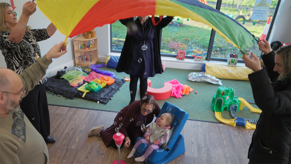 A group of adults holding a colourful parachute above a small child sat in a chair