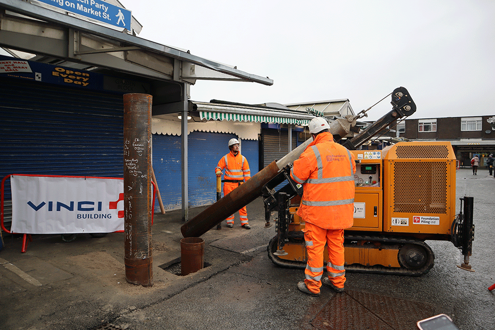 Two people in high-visibility clothing putting a large pipe into a hole in the ground
