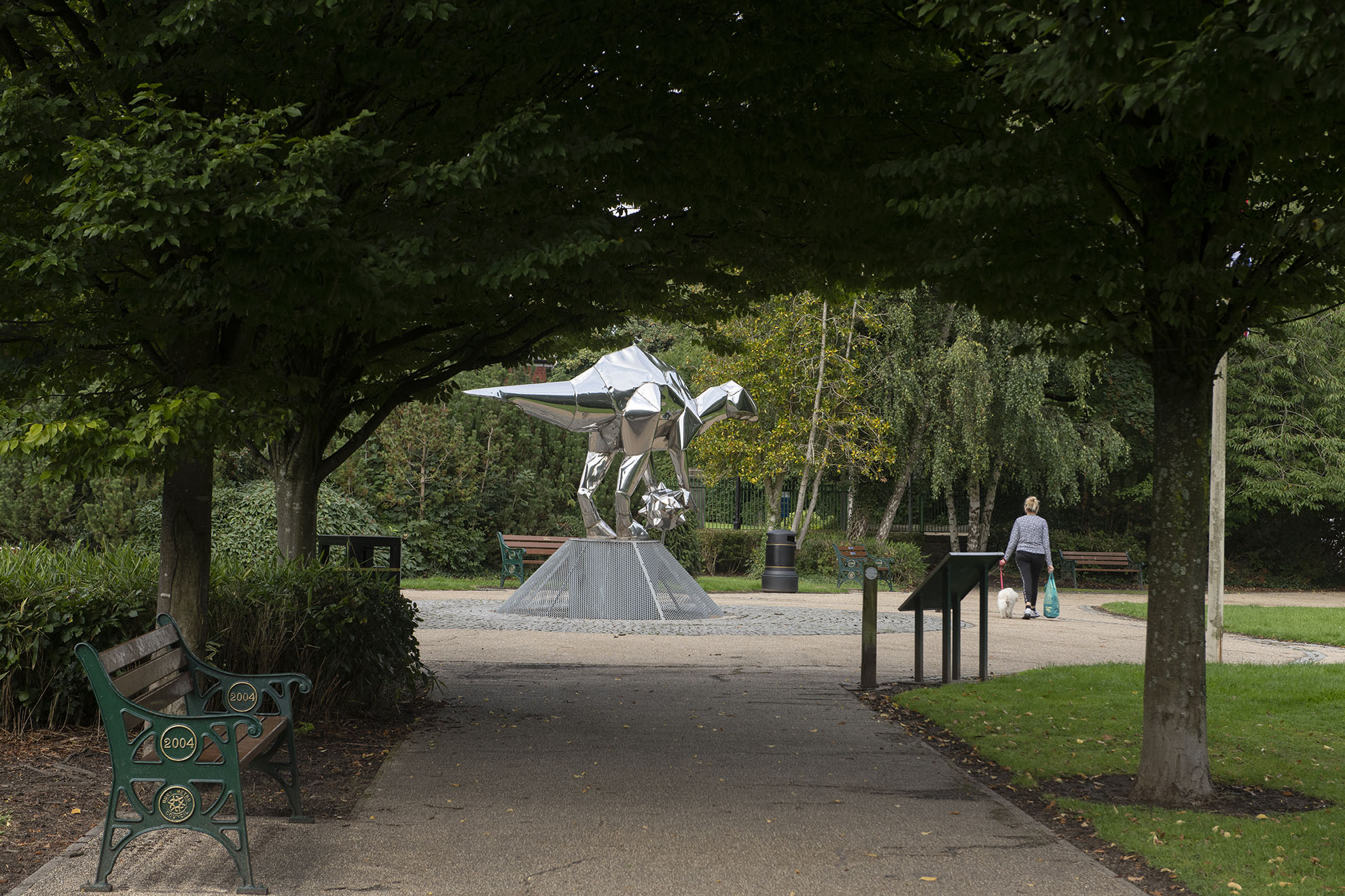 A pathway with a bench on the left leading towards a large polished steel sculpture shaped as a dragon
