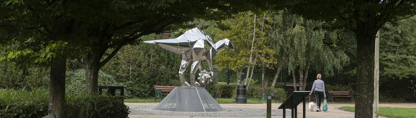 A pathway with a bench on the left leading towards a large polished steel sculpture shaped as a dragon