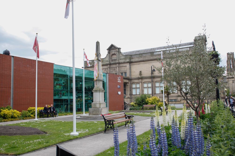 A brick building surrounded by gardens, plants with a war memorial in the centre.