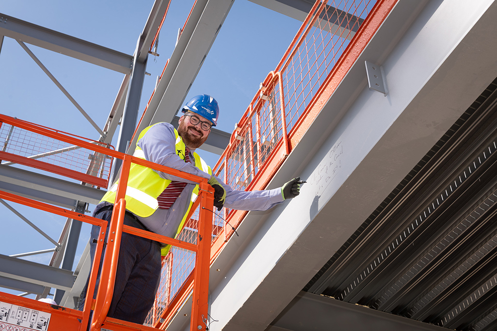 A person wearing protective clothing who is high up in a building on a construction site and pointing to a beam