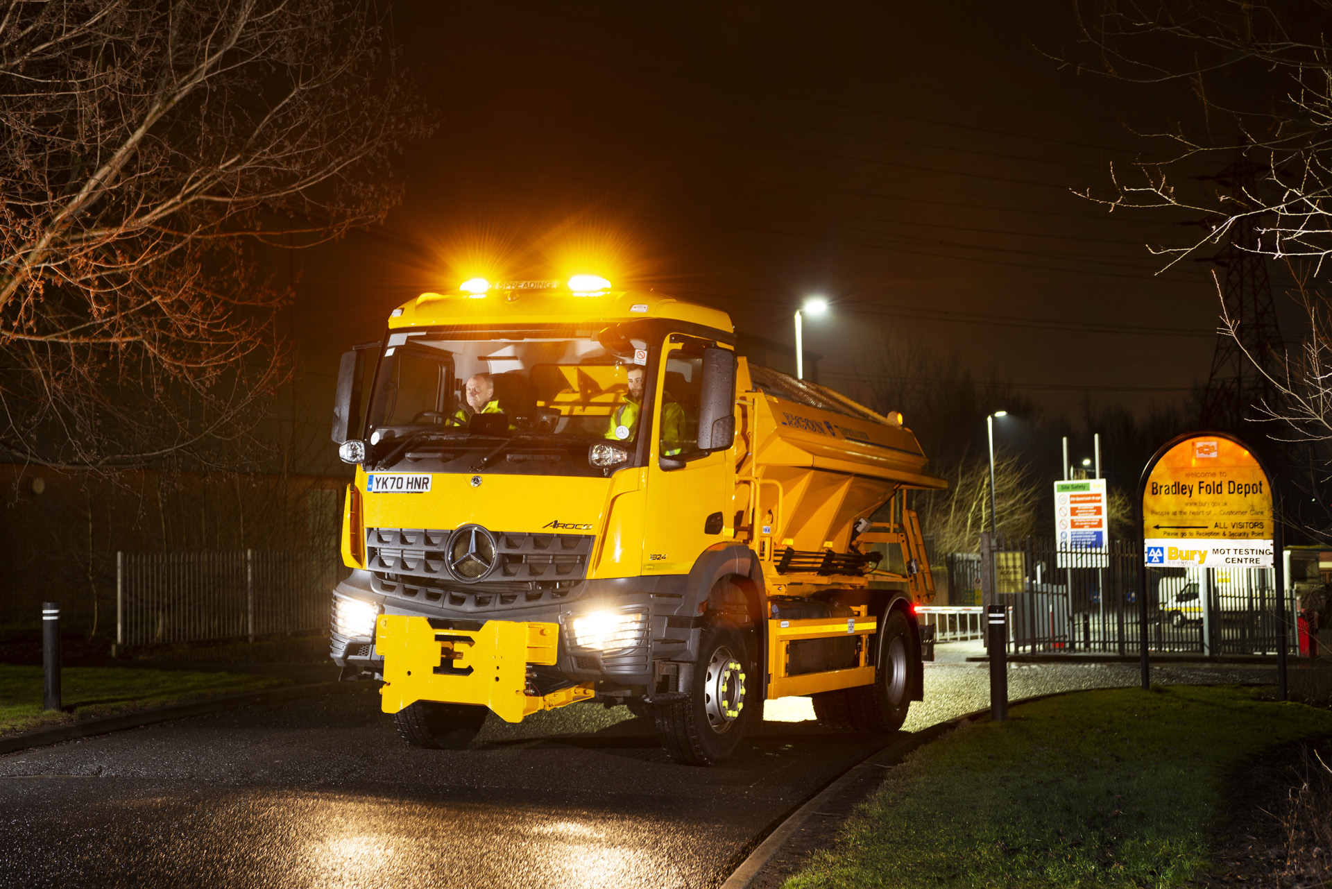 A large yellow vehicle travelling on a road in the dark with the headlights on