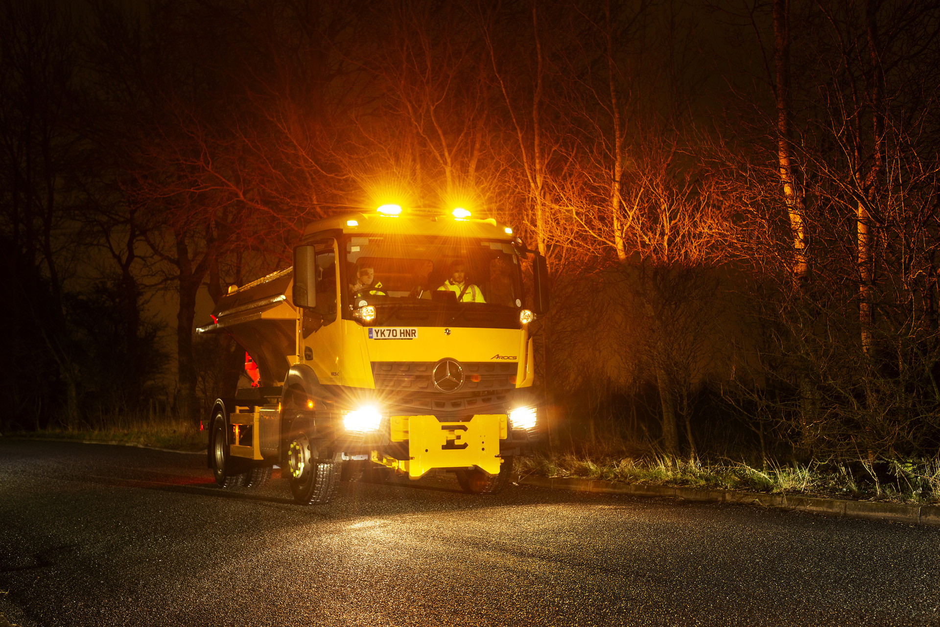 A large yellow vehicle travelling on a road in the dark with the headlights on