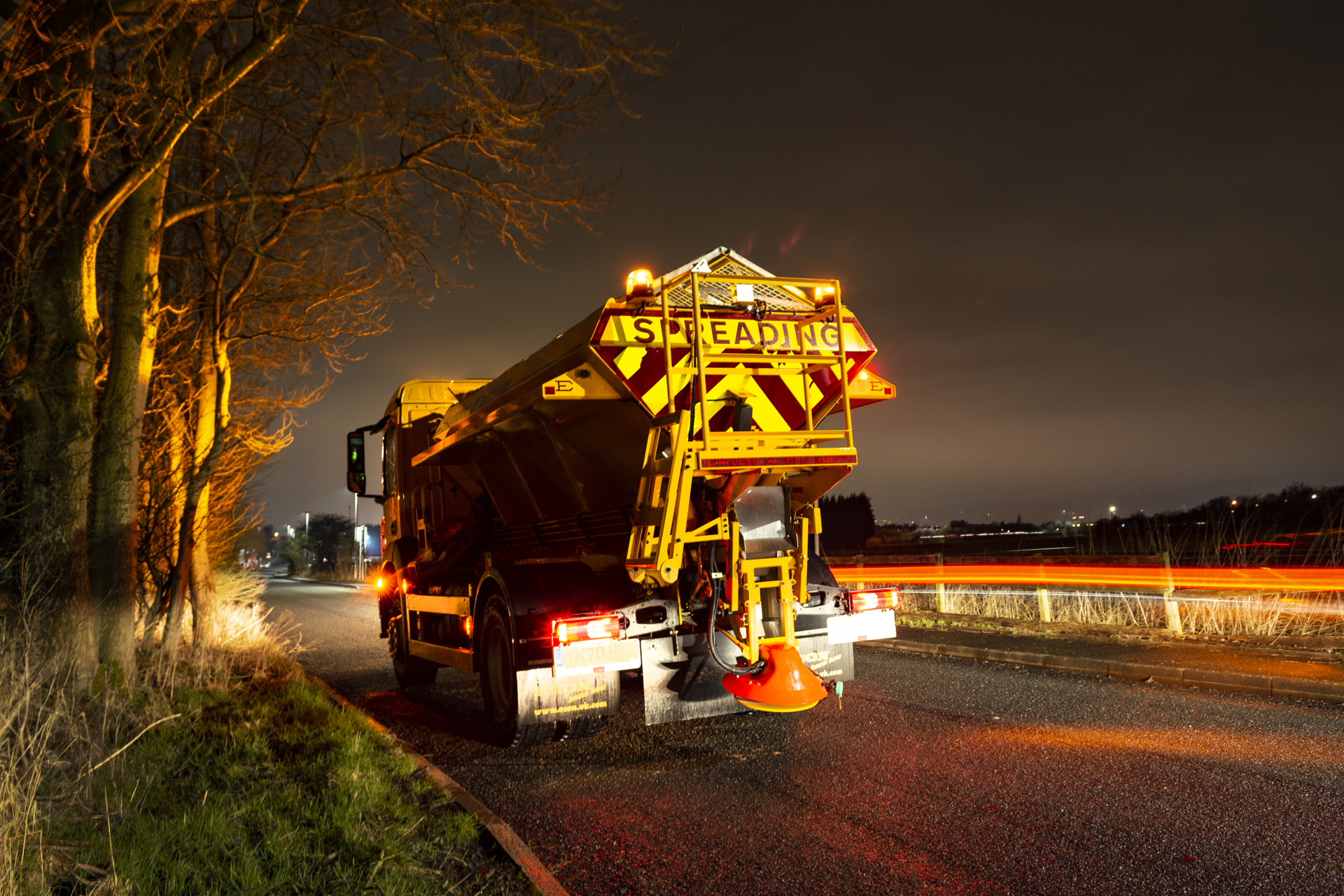 A large truck, with the word 'spreading' written on the back, travelling on a road in the dark