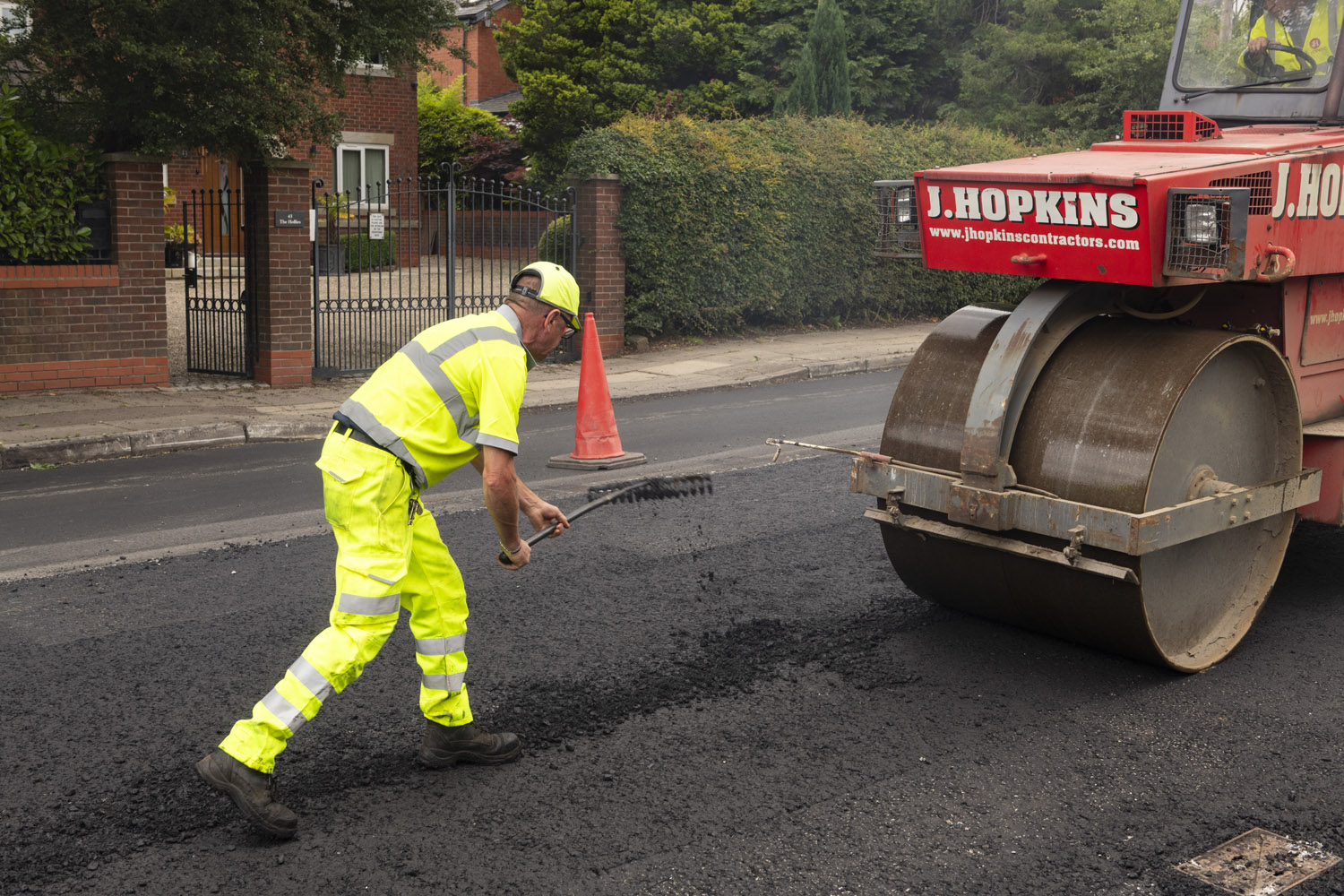A person wearing high-visibility clothes in front of a machine that is rolling along a road surface