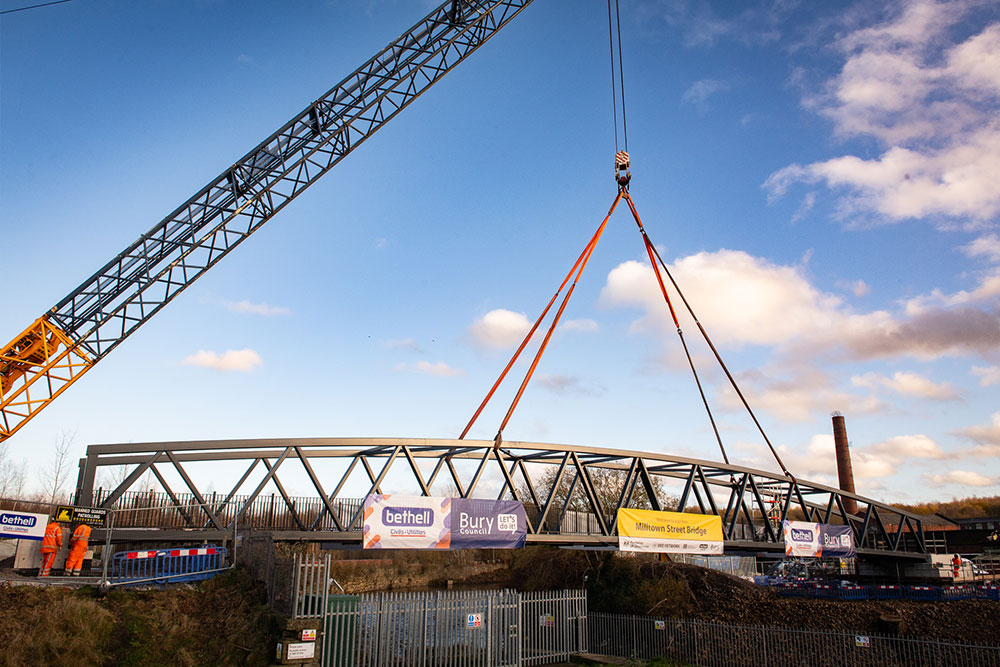 A crane lifts a curved steel pedestrian bridge over a river, with advertising banners attached and a clear blue sky in the background.