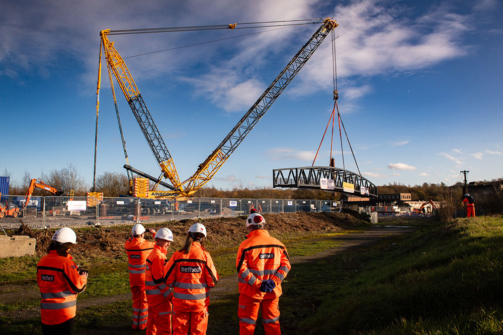 A group of engineers in high-visibility orange safety clothing and white helmets stand in the foreground watching as a large crane lifts a steel pedestrian bridge over a river