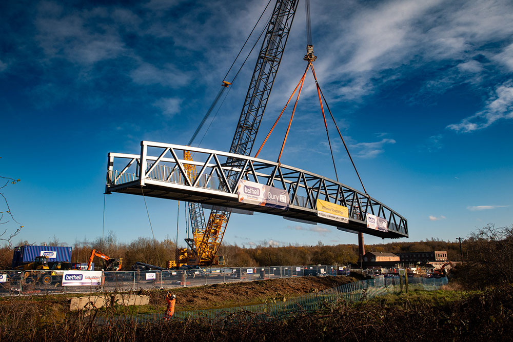 A large crane lifts a curved steel pedestrian bridge over a river, with advertising banners attached and a clear blue sky in the background.