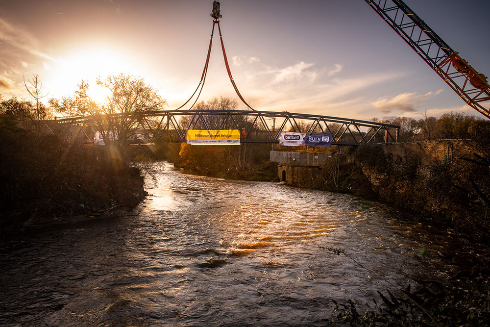 A crane suspends a curved steel pedestrian bridge over a flowing river at sunset, with lifting straps visible above the deck