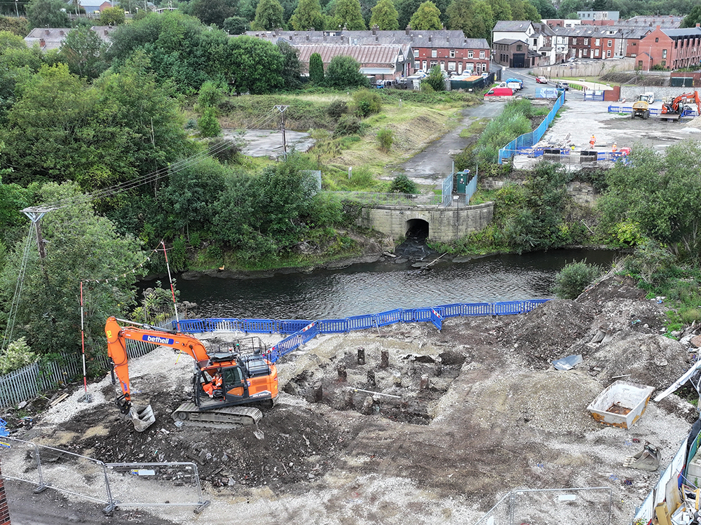 A river with a building site on one side and houses and a road on the other side