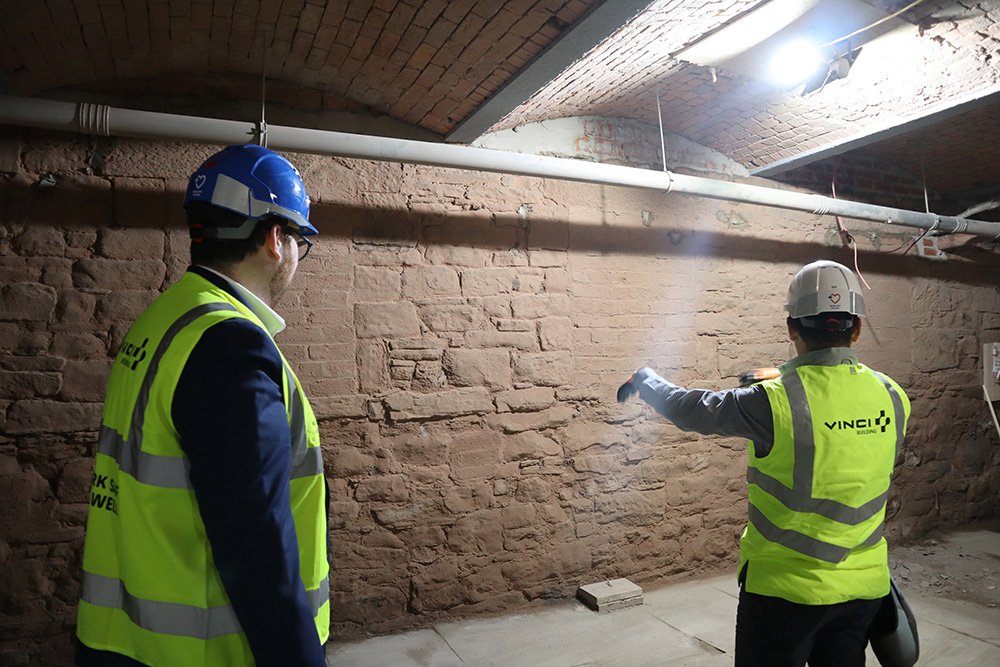 Two people wearing safety clothing, in a building basement, inspecting a wall