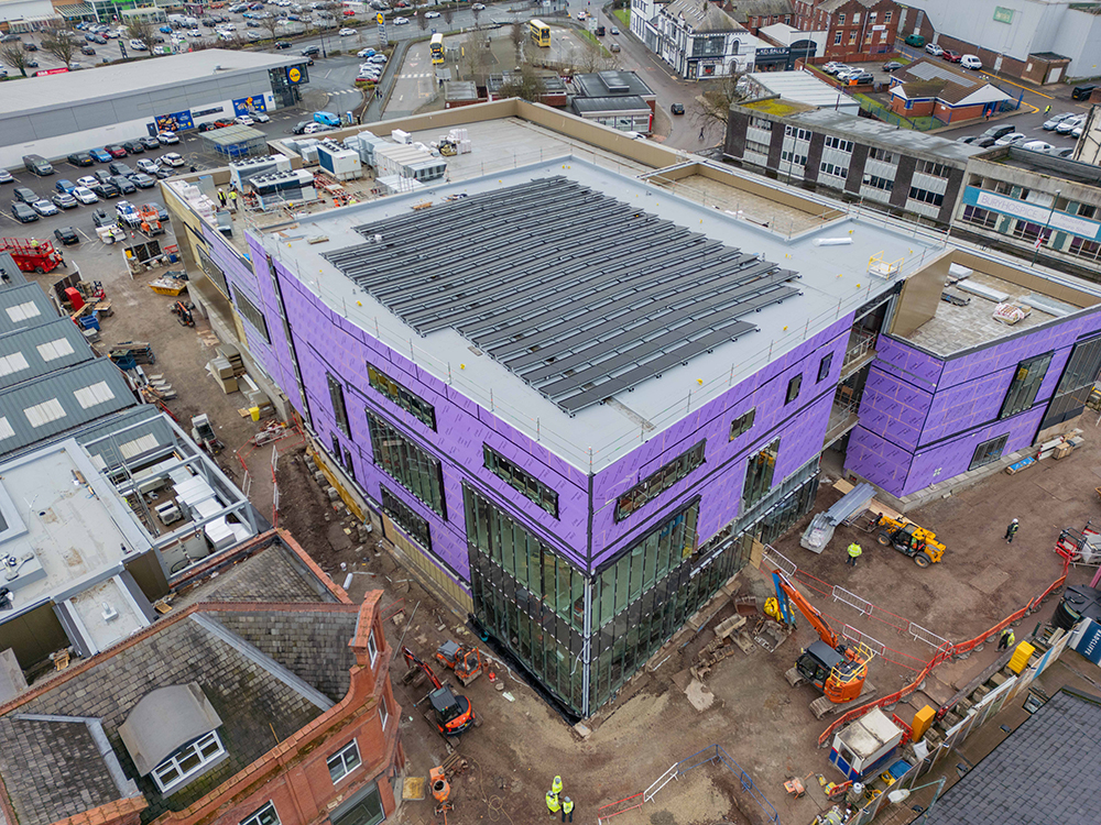 A birds-eye view of a large building on a construction site surrounded by other large buildings and a car park
