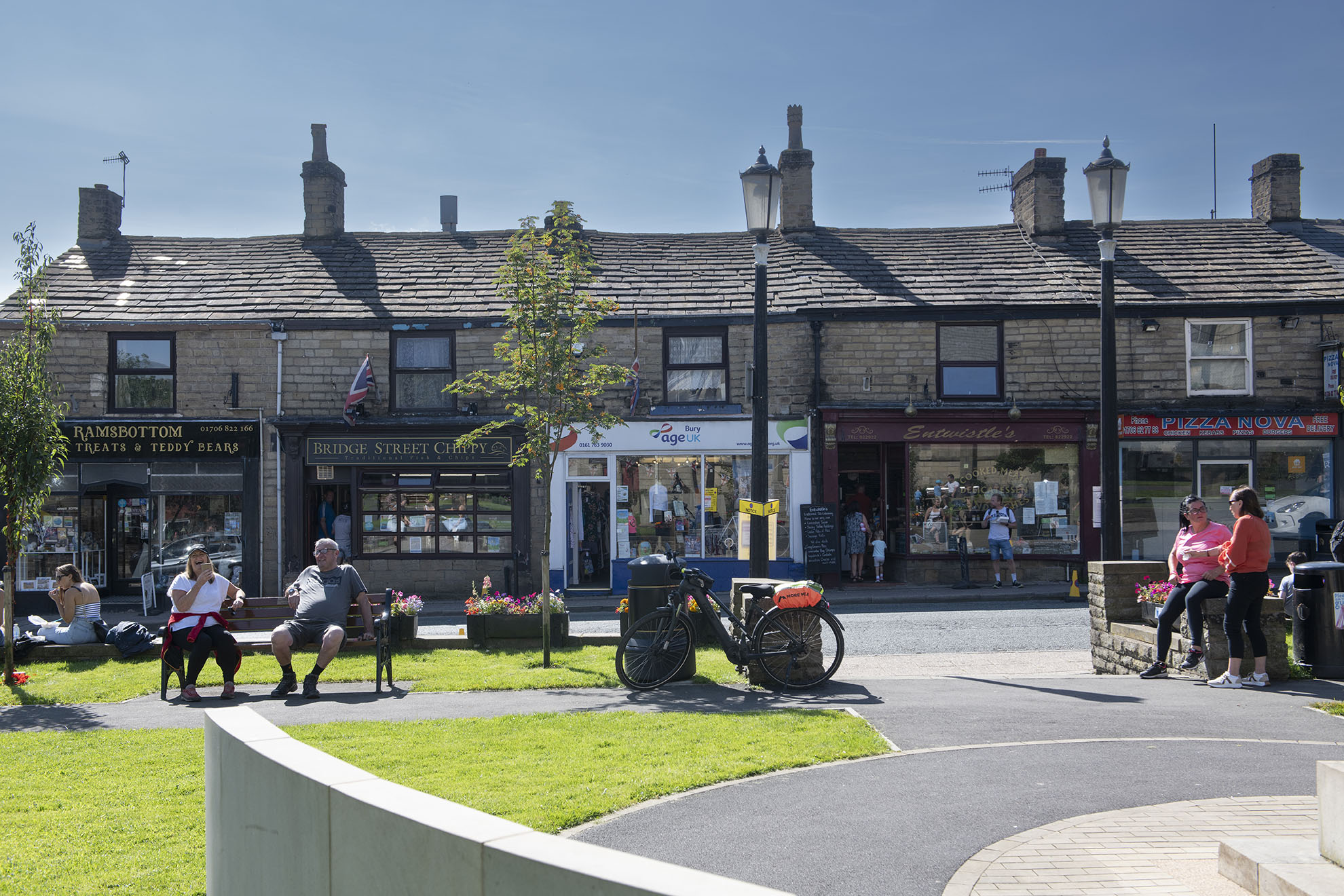Row of small stone-fronted shops on a sunny day, with people sitting on benches outside and others standing nearby; a bicycle is parked by a lamp post in the foreground.