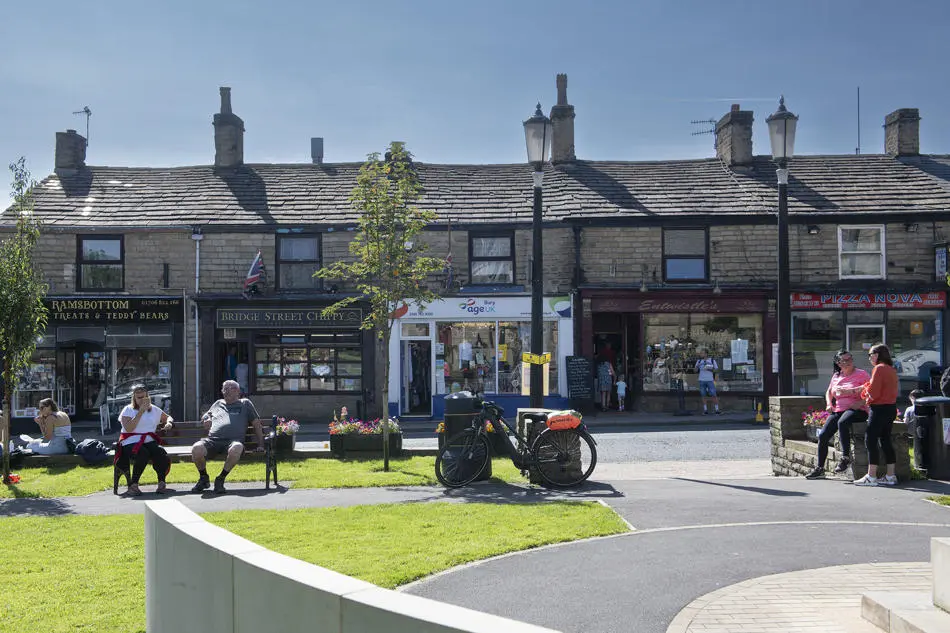 Row of small stone-fronted shops on a sunny day, with people sitting on benches outside and others standing nearby; a bicycle is parked by a lamp post in the foreground.